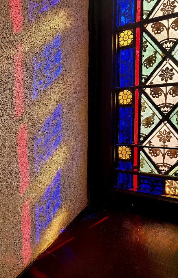 Stained glass window casts colorful light patterns on a textured wall, showcasing intricate designs and vibrant hues in a historical setting.