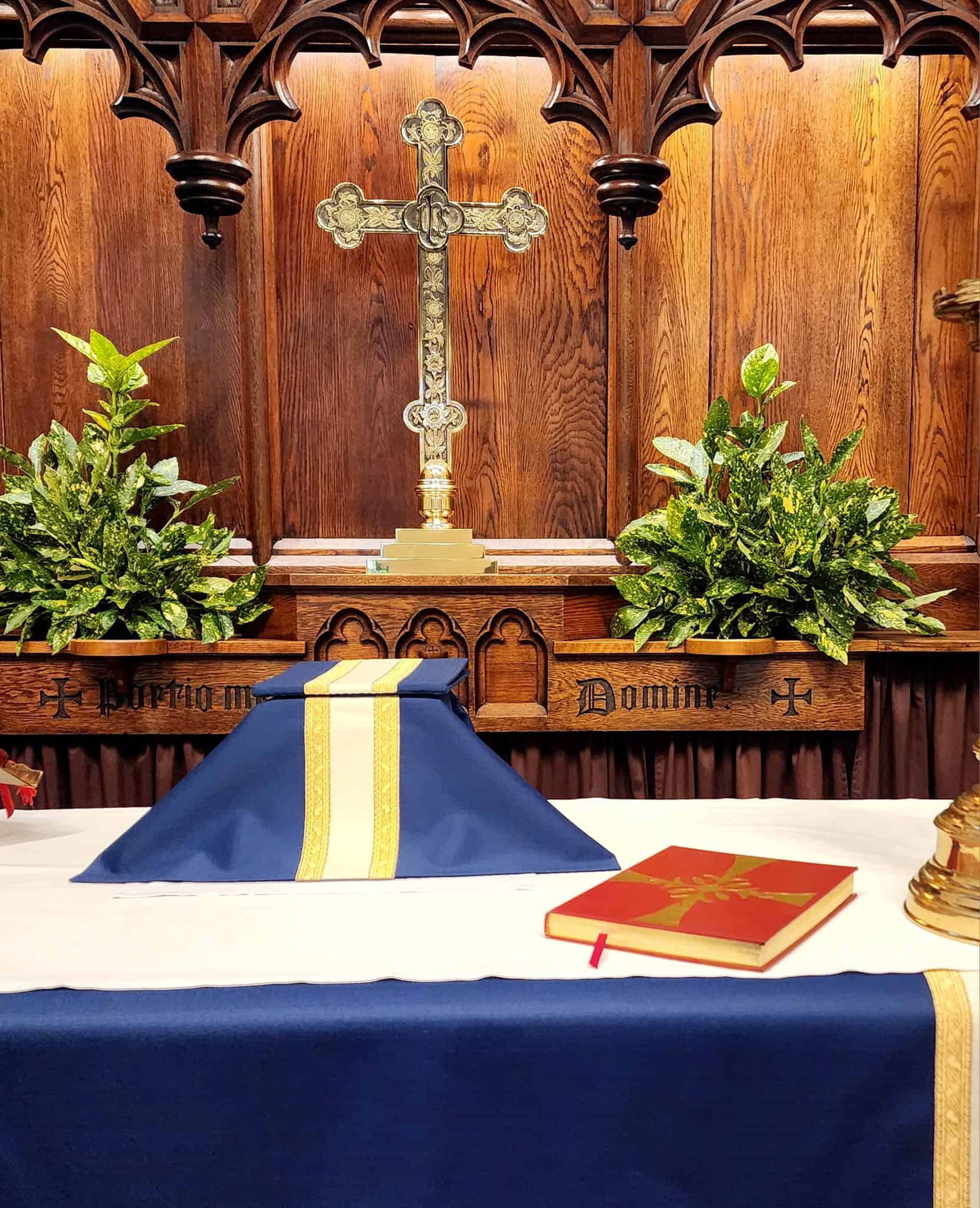An ornate altar featuring a cross, surrounded by greenery, with a book resting on a table adorned in blue and gold fabric.