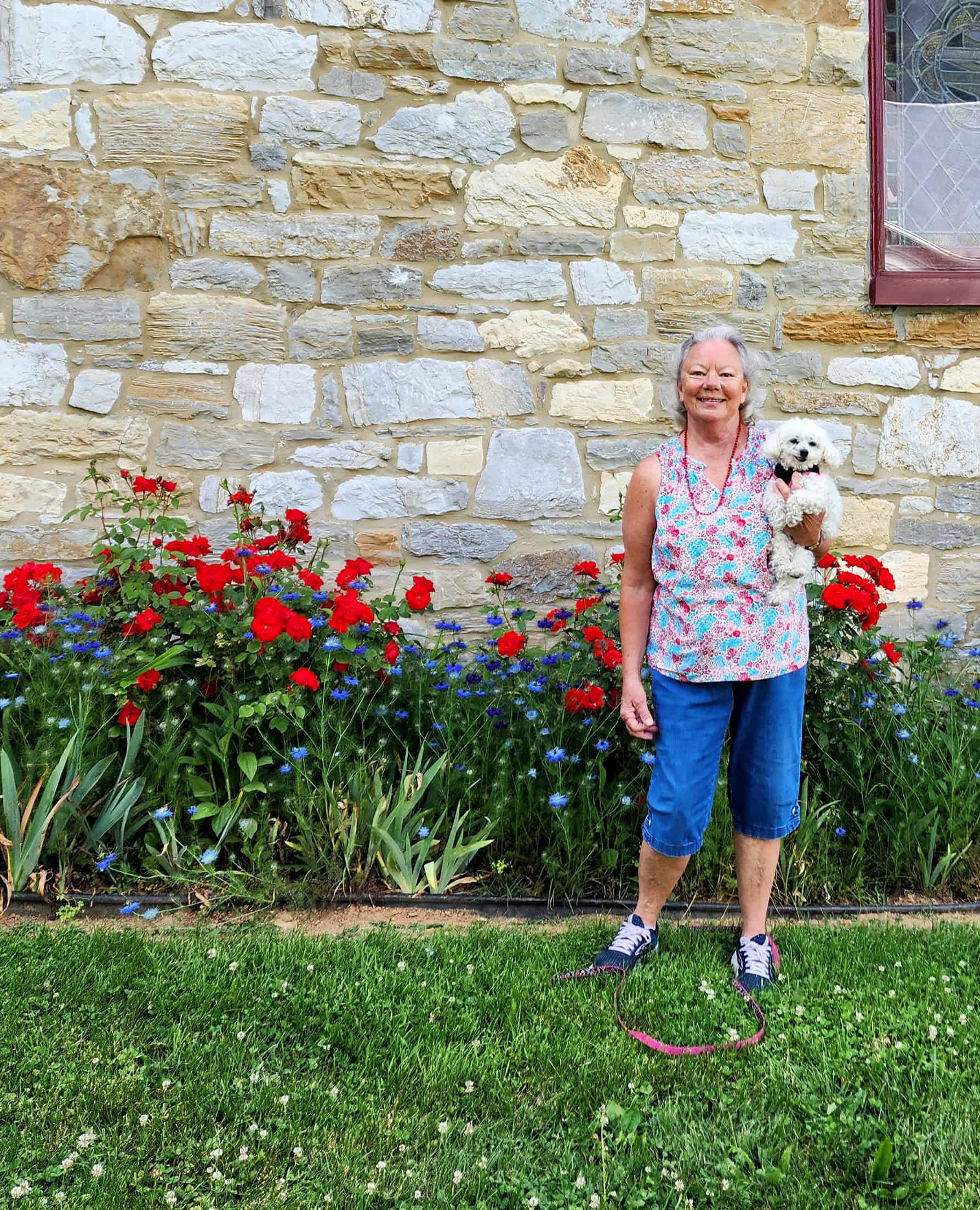 A person wearing a visor and gloves tends a vibrant garden of pink and white flowers, with trees and a water tower visible behind.