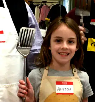 A child named Alyssa holds a spatula, standing between two people in aprons, likely participating in a cooking or kitchen activity.