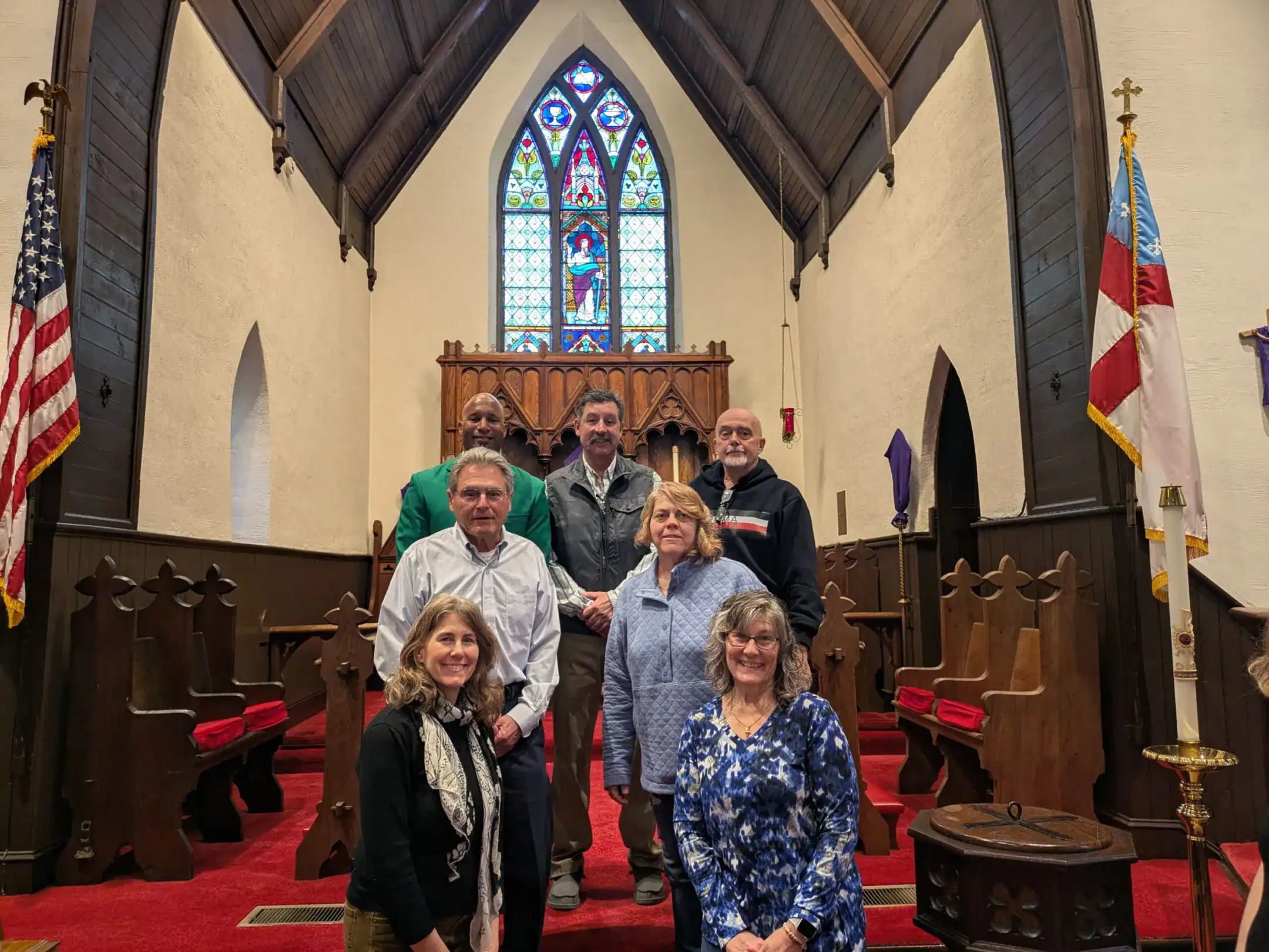 Seven people stand inside a church with stained glass windows and flags, featuring wooden pews and an elevated altar area.